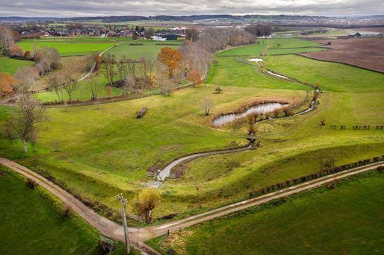 Dronebeeld van overstromingszone: de waterloop kronkelt door het landschap. De dijk ligt dwars op de waterloop.